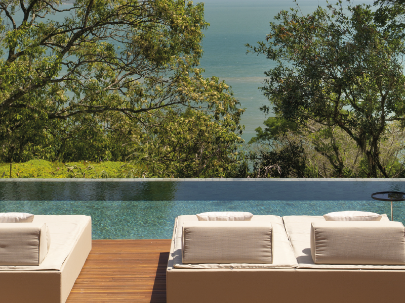 Espreguiçadeiras junto a uma piscina infinita turquesa com vista para as árvores e um lago tranquilo.