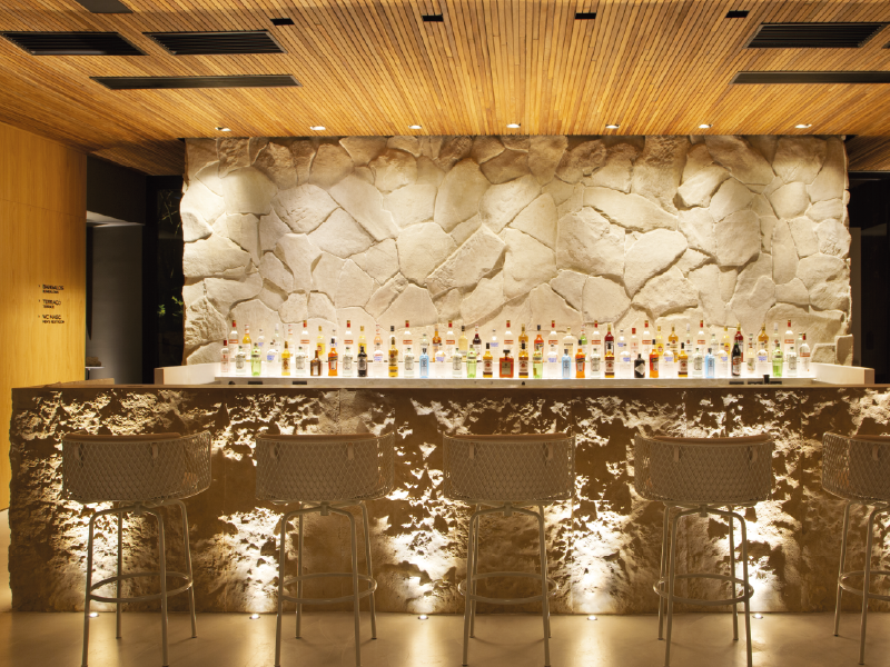 Bar moderno com parede de pedra, teto de madeira, balcão retroiluminado e fileiras de garrafas.