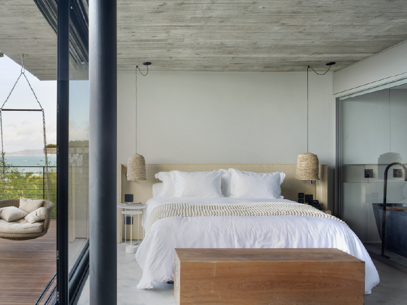 Quarto minimalista com cama branca, cadeiras de vime e vista para o mar a partir do deck.