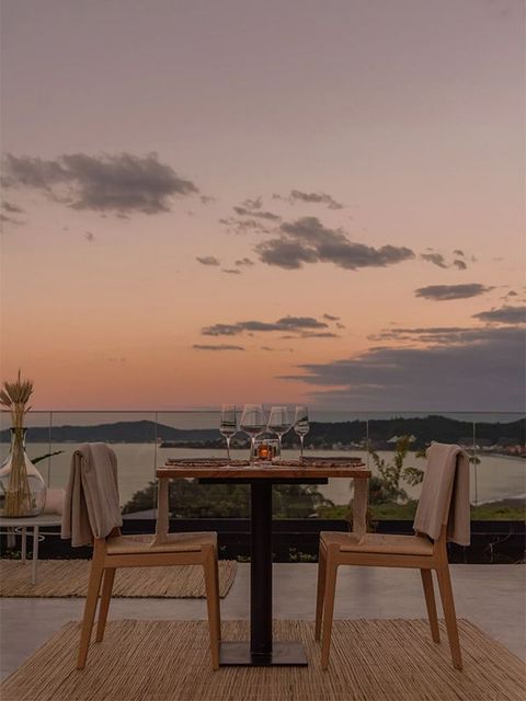 Mesa de jantar com copos e cadeiras com vista para o mar ao pôr do sol.