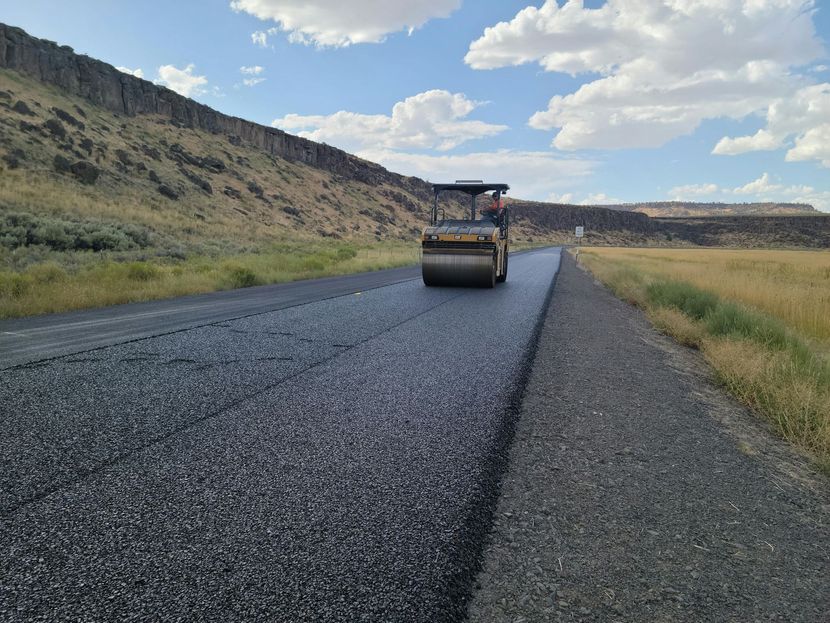 A construction vehicle rolls over newly laid, dark asphalt on a rural road alongside a rocky hillside under a cloudy sky.