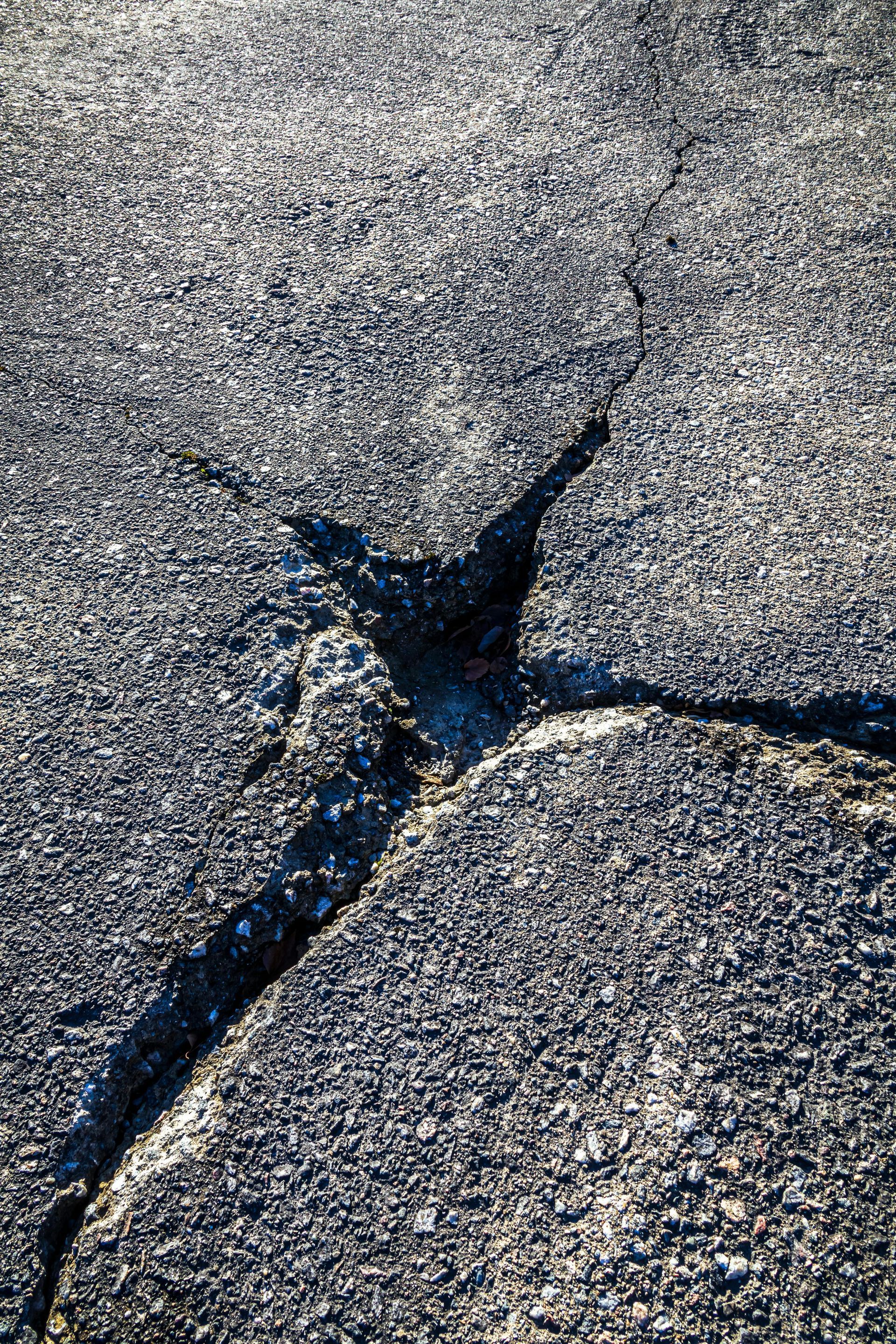 A deep, jagged crack in an asphalt surface spreading out from a central point, illuminated by sunlight.