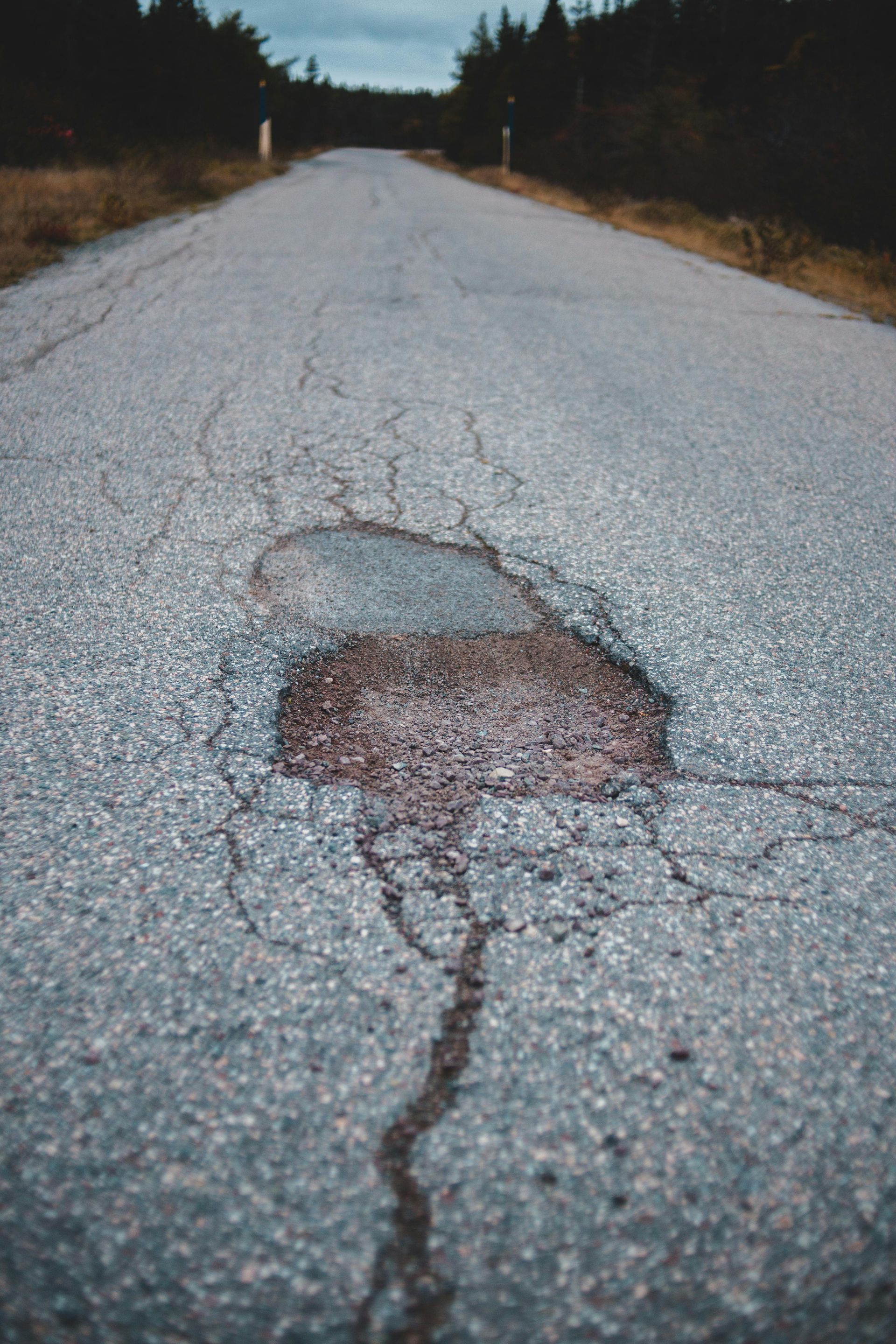 A weathered, grey asphalt road stretches toward a distant forest, featuring a prominent, cracked pothole in the foreground.