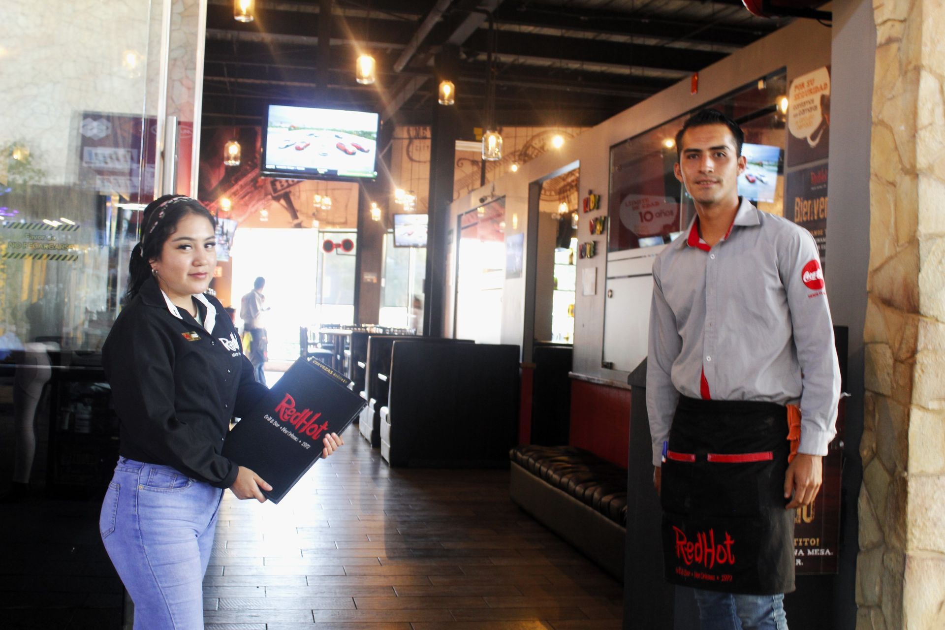 Dos empleados del restaurante de pie en el interior, sosteniendo menús negros en un comedor con poca luz.