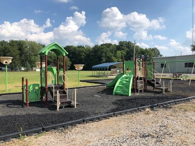 A playground with green equipment, slides, and swings set on a dark rubber mulch surface under a partly cloudy sky.