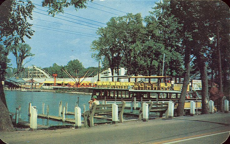 A vintage postcard shows a riverboat docked at a pier, with an amusement park featuring a roller coaster in the background.
