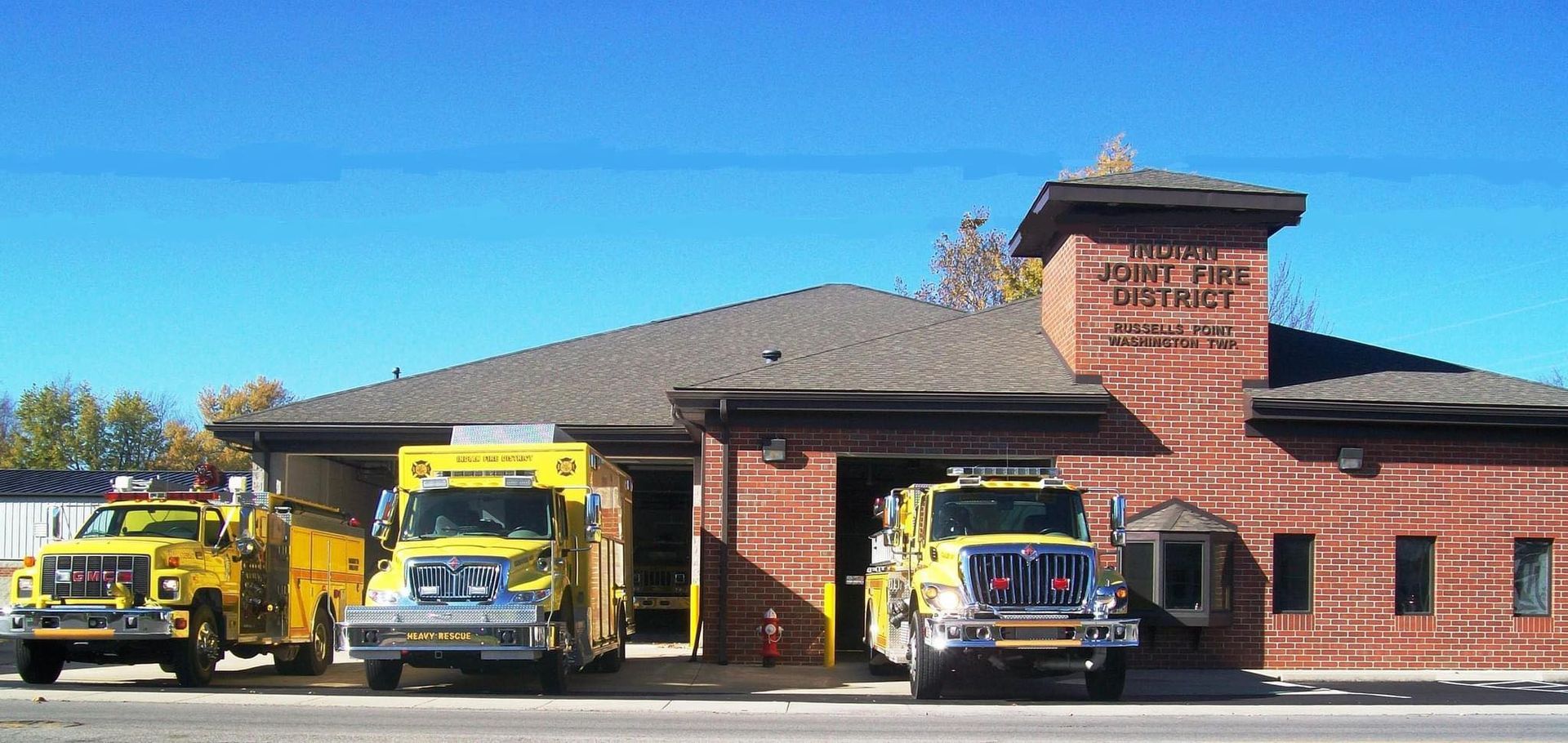 Three yellow emergency vehicles parked in front of a brick fire station building on a clear, sunny day.