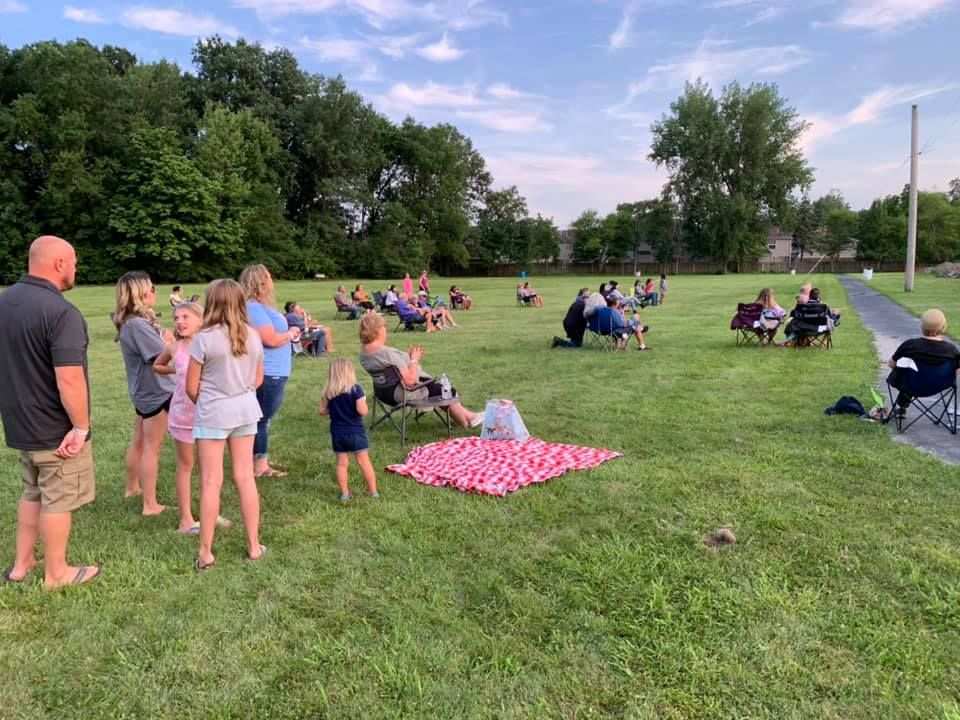 People gather on a grassy field in folding chairs and on blankets for an outdoor event under a clear, sunny sky.