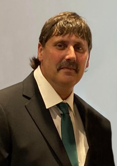 A person with short brown hair and a mustache wearing a black suit, white collared shirt, and a dark teal tie.