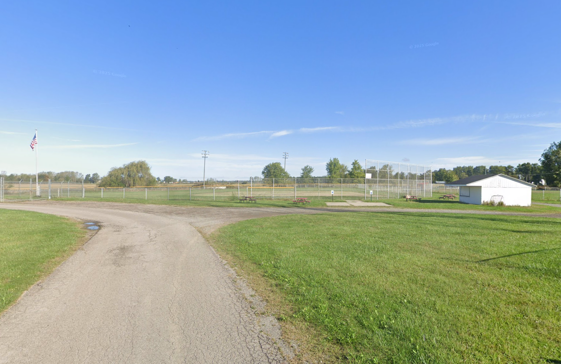 A dirt path leads toward a large, grassy field with a fenced sports area and a small white building under a clear blue sky.