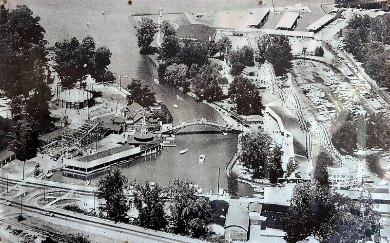 Aerial view of an amusement park with a river, a bridge, rides, and surrounding trees in black and white.