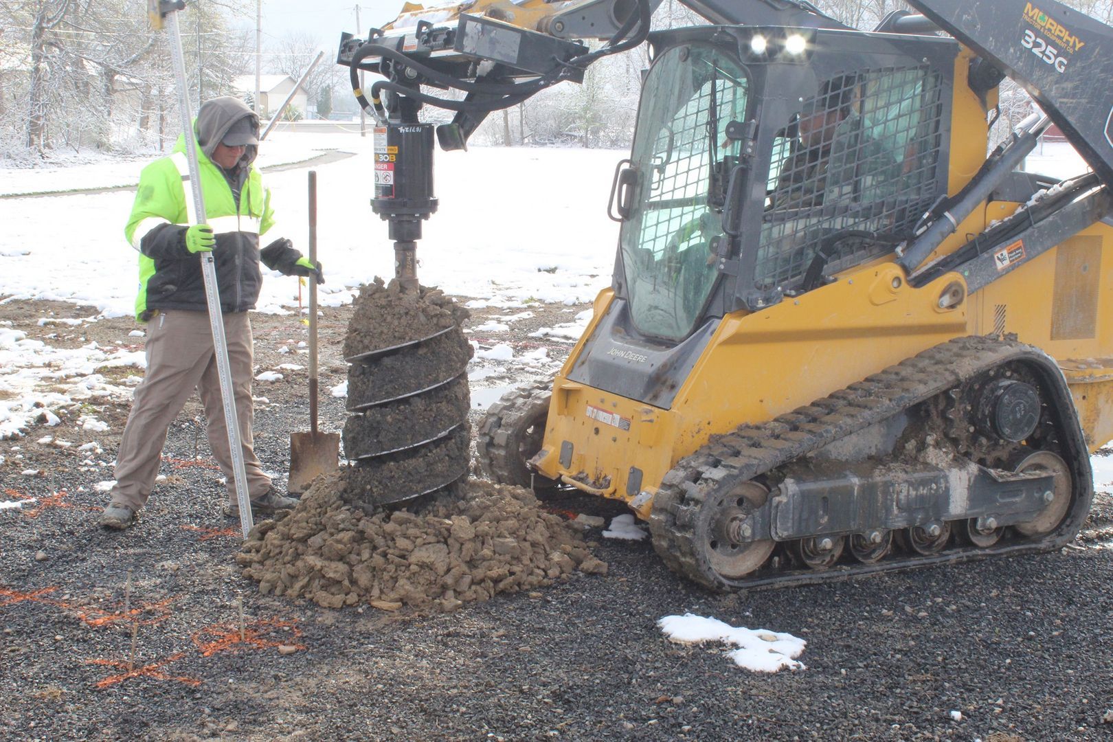A person in a neon yellow jacket uses a skid steer with an auger attachment to drill a hole in a snowy, gravel-covered lot.