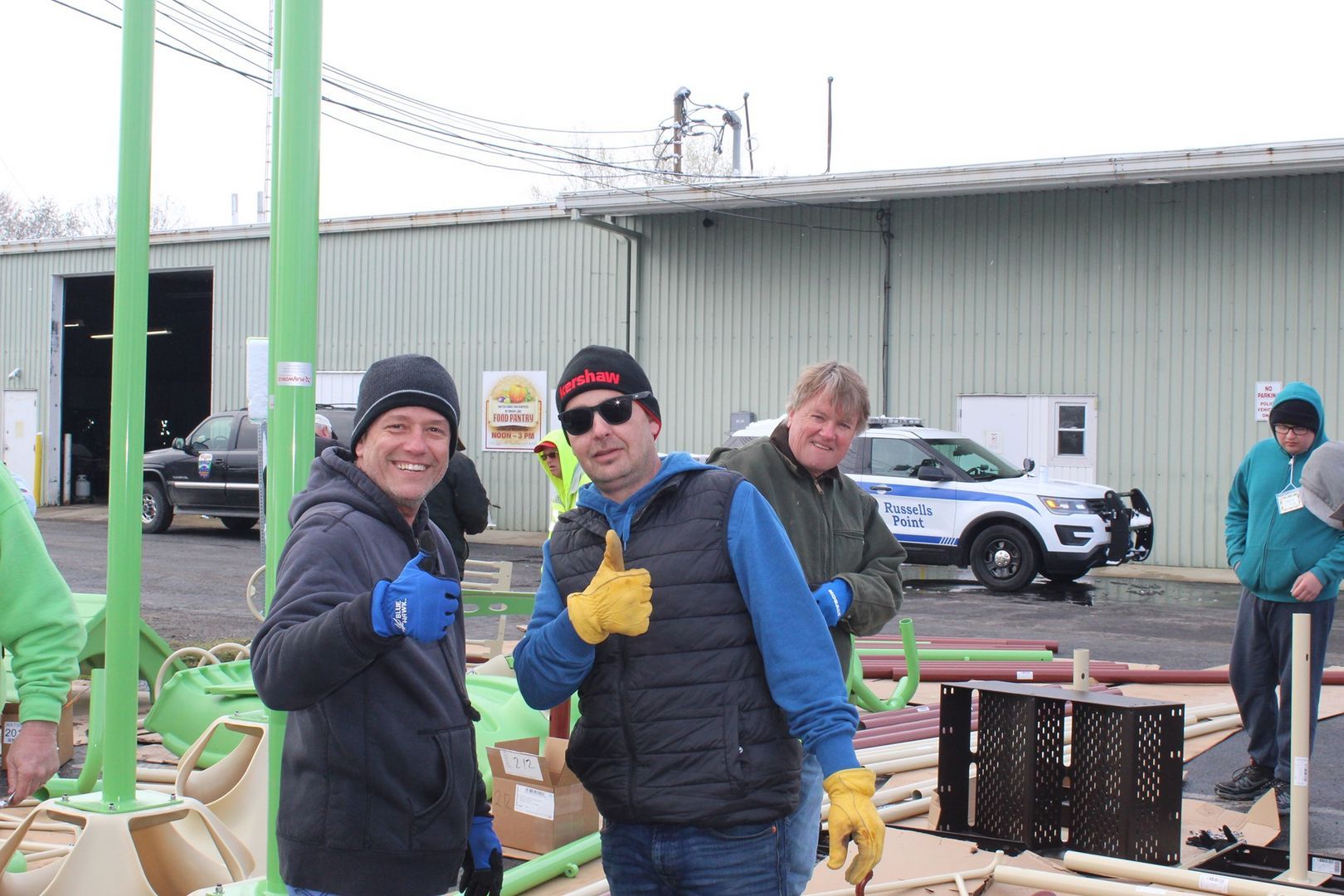 Three people smile and give thumbs-up while assembling playground equipment in an outdoor industrial lot.