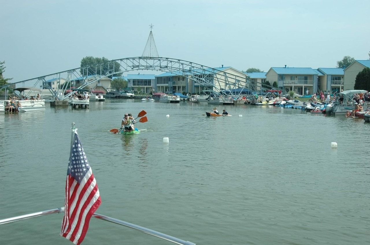 An American flag flies in the foreground as people kayak on a canal lined with blue-roofed buildings and a large bridge.