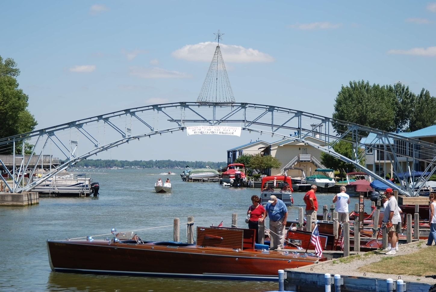 A classic boat docked at a marina under a decorative blue arched footbridge on a sunny day.
