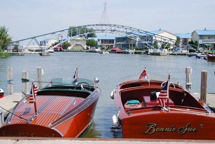 Two polished wooden motorboats docked side-by-side at a harbor in front of a metal arch bridge.