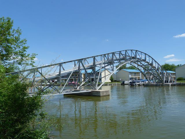 A metal arch pedestrian bridge spans a calm body of water on a sunny day, with houses visible in the background.