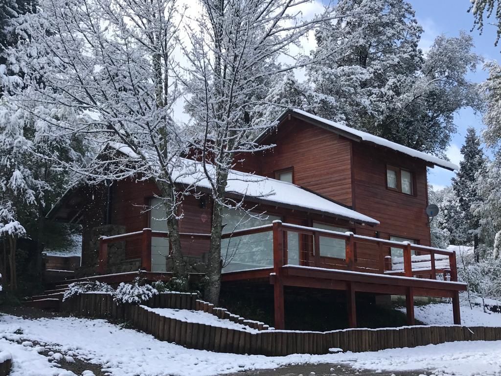 Una cabaña rústica de madera de dos pisos se encuentra rodeada de árboles cubiertos de nieve y una ligera capa de nieve en el suelo.