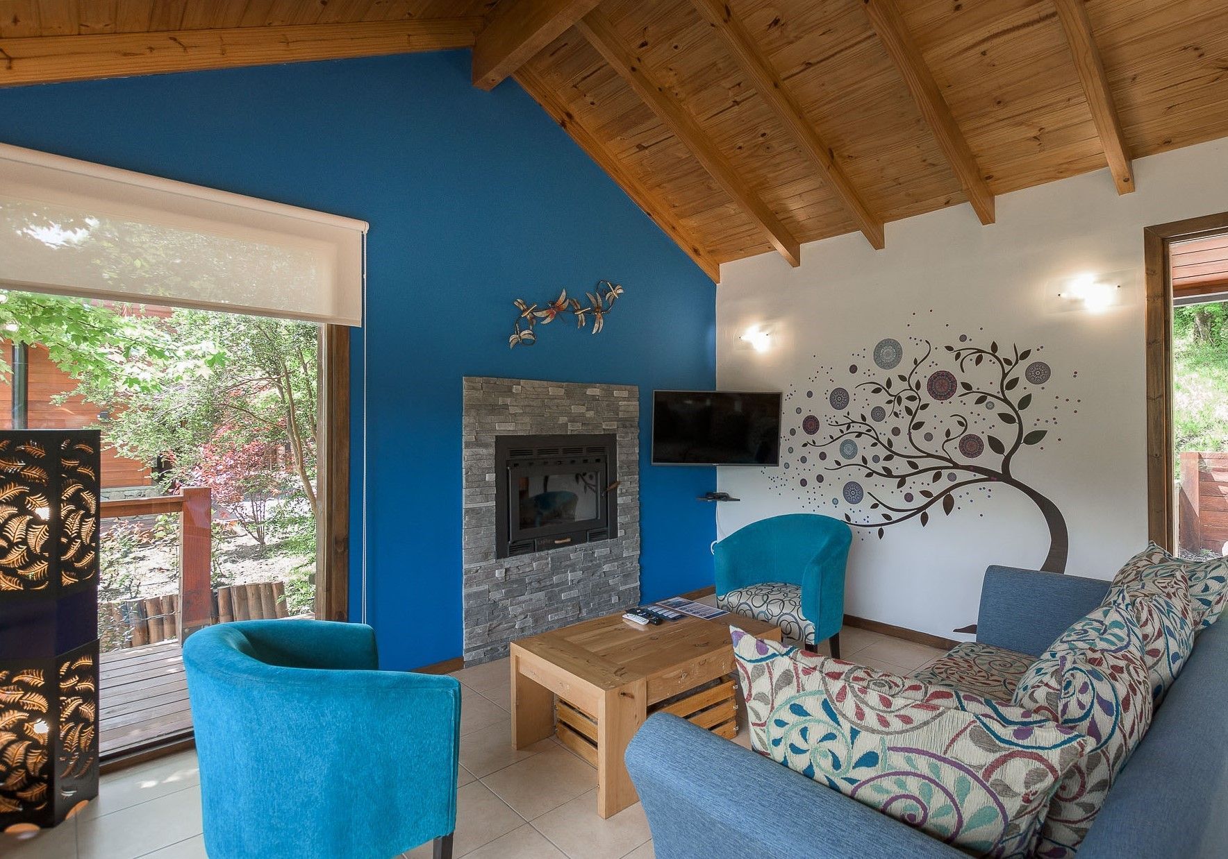 Una sala de estar con paredes decoradas en azul, techo con vigas de madera, chimenea de piedra, sillones azules modernos y un sofá con estampado floral.