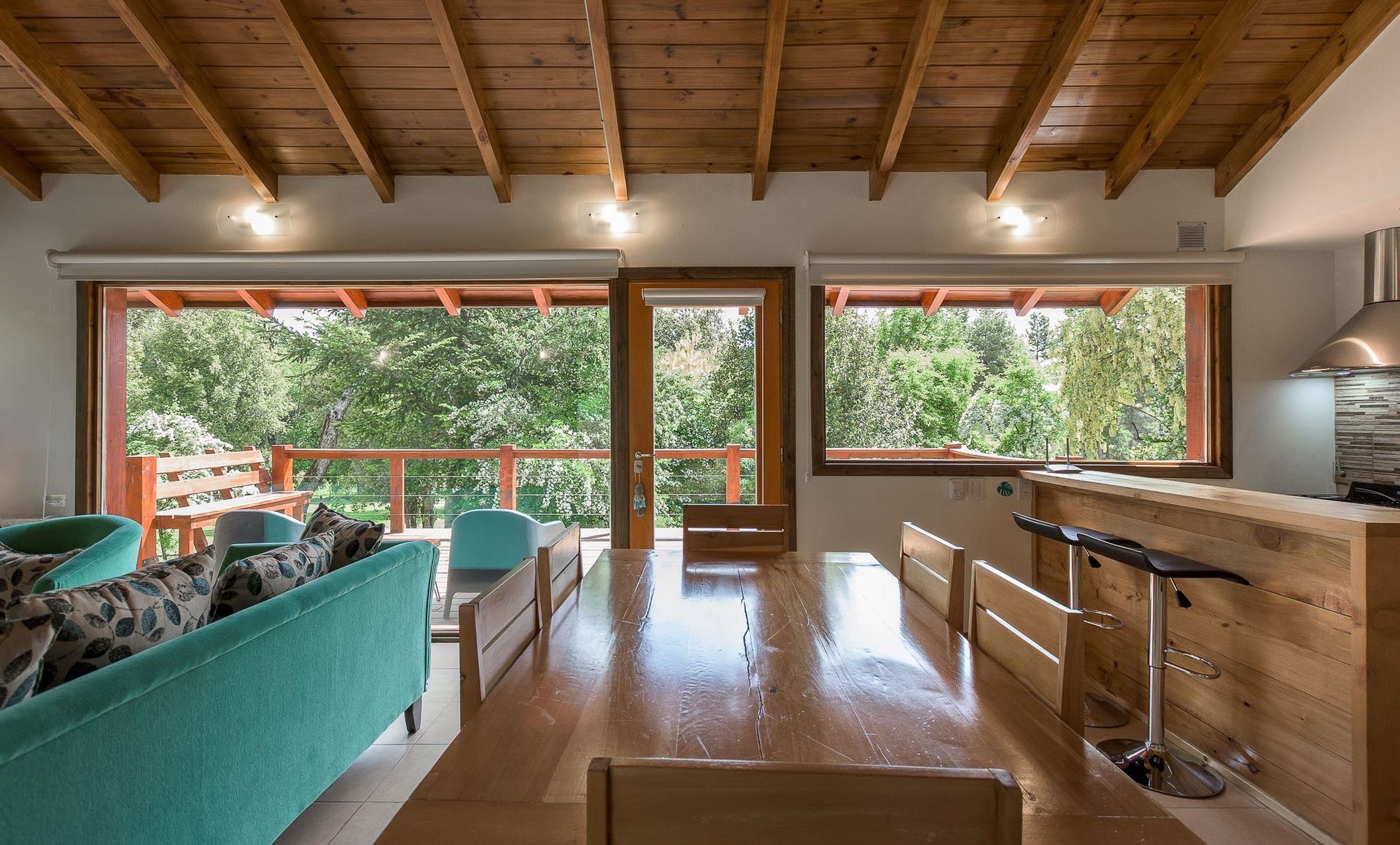Una luminosa sala de estar diáfana con un sofá color turquesa, una mesa de comedor de madera y una barra de desayuno con vistas a una terraza arbolada.