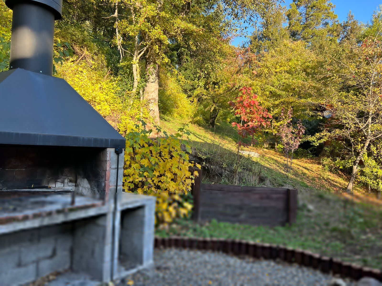 Una chimenea exterior de piedra gris se alza en un patio de grava, con vistas a una ladera cubierta de árboles otoñales.