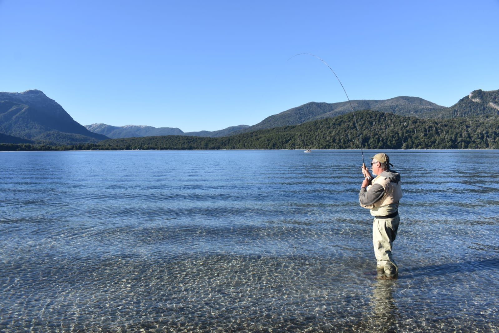 Una persona con botas de agua pesca con mosca en un lago tranquilo y cristalino rodeado de montañas boscosas bajo un cielo azul.