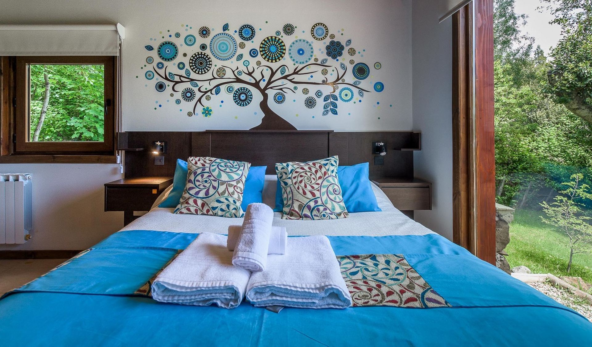 Un dormitorio con una cama con funda azul, almohadas estampadas, toallas blancas dobladas y un mural decorativo con un árbol en la pared.