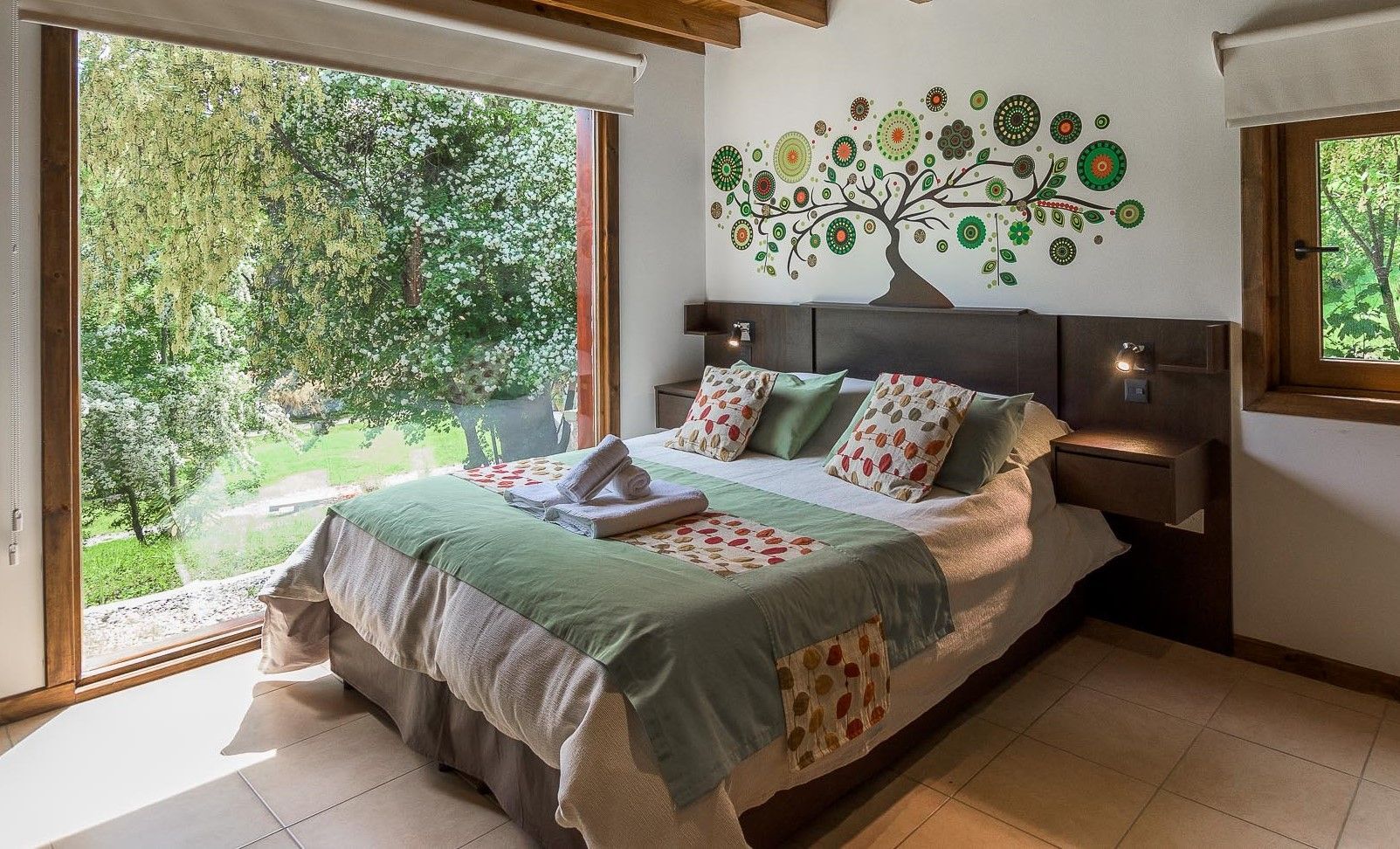 Un dormitorio con una cama con ropa de cama estampada, un mural en la pared que representa un árbol y una gran ventana con vistas a una exuberante vegetación.