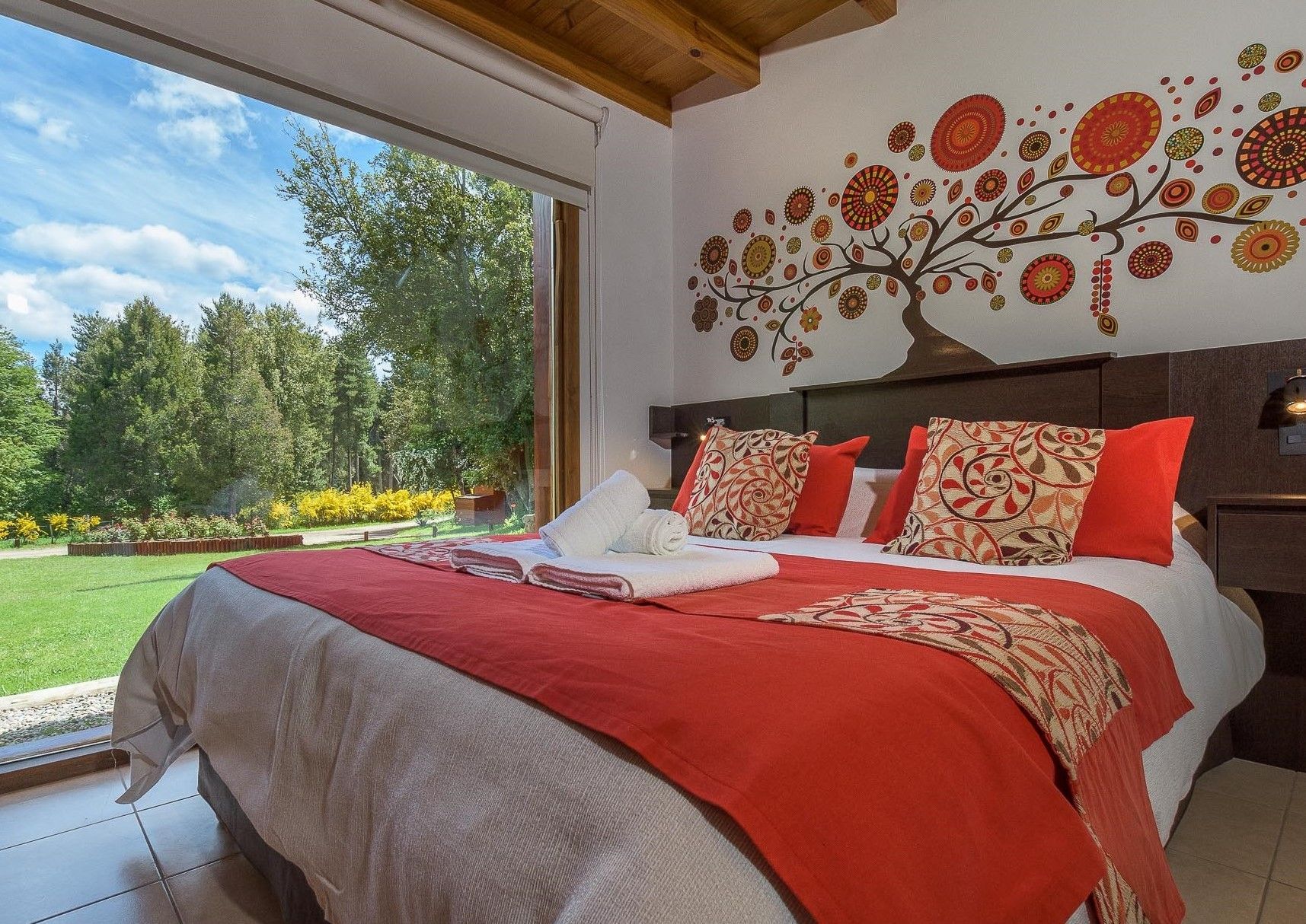 Una habitación de hotel con una cama grande, ropa de cama naranja y un mural floral en la pared, con vistas a un jardín exuberante.