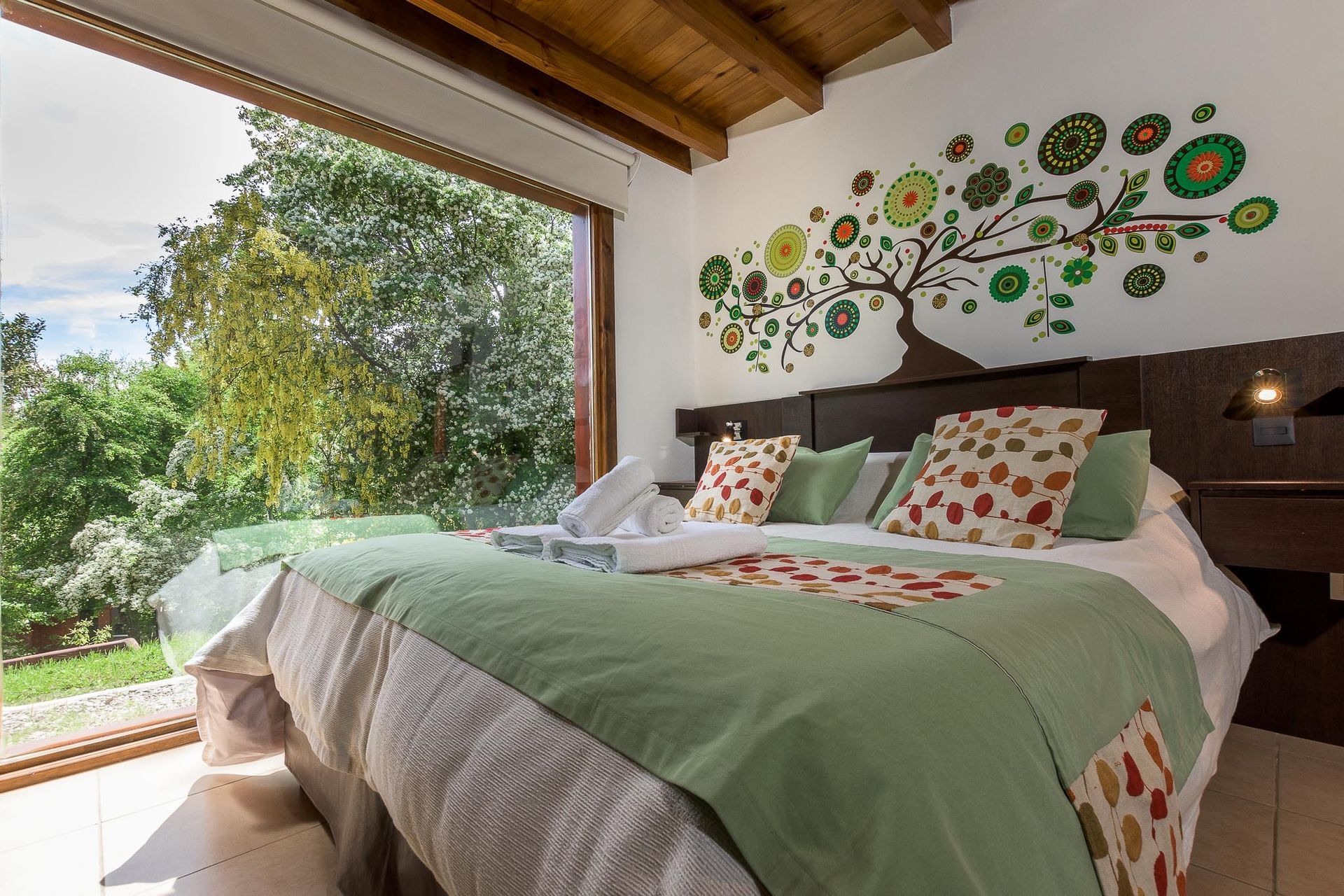 Un dormitorio moderno con una cama con ropa de cama verde y floral, situado contra una pared con un mural de un árbol y una gran ventana.
