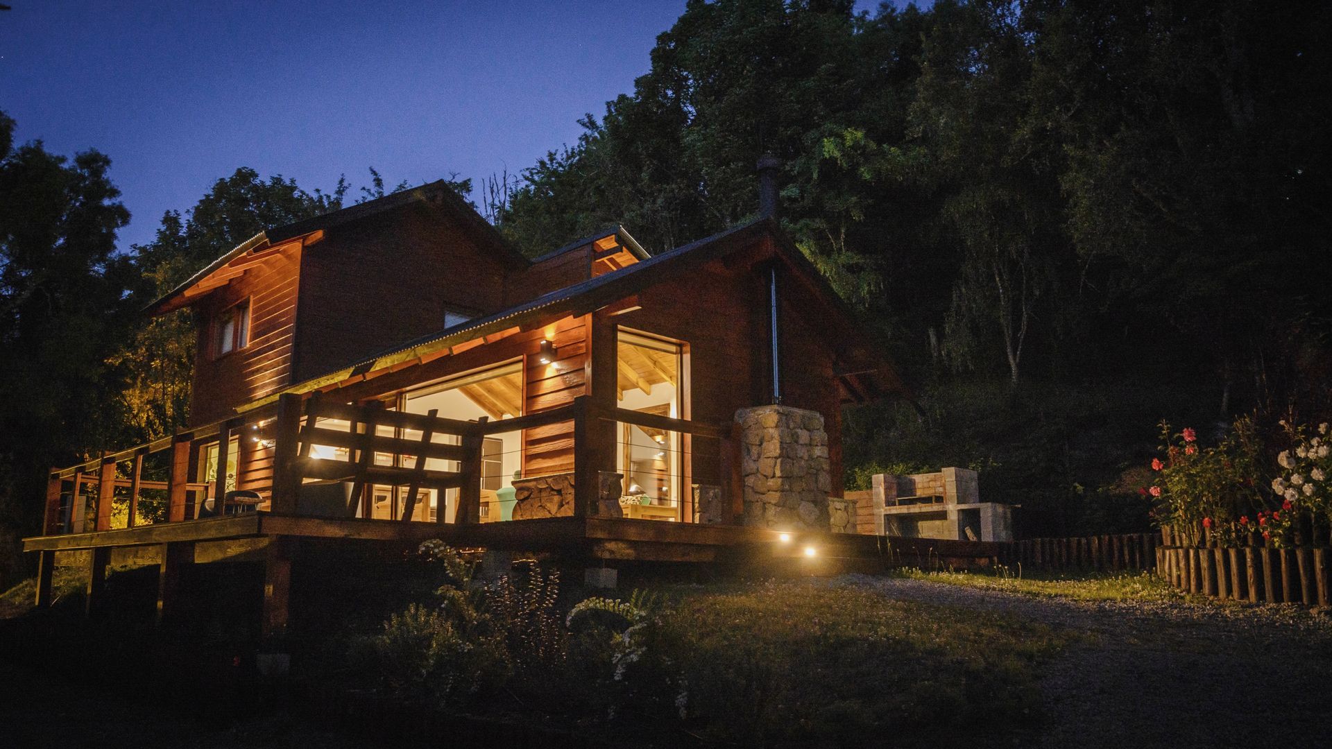 Una acogedora cabaña de madera iluminada, rodeada de árboles al anochecer, con una terraza de madera y una chimenea de piedra.
