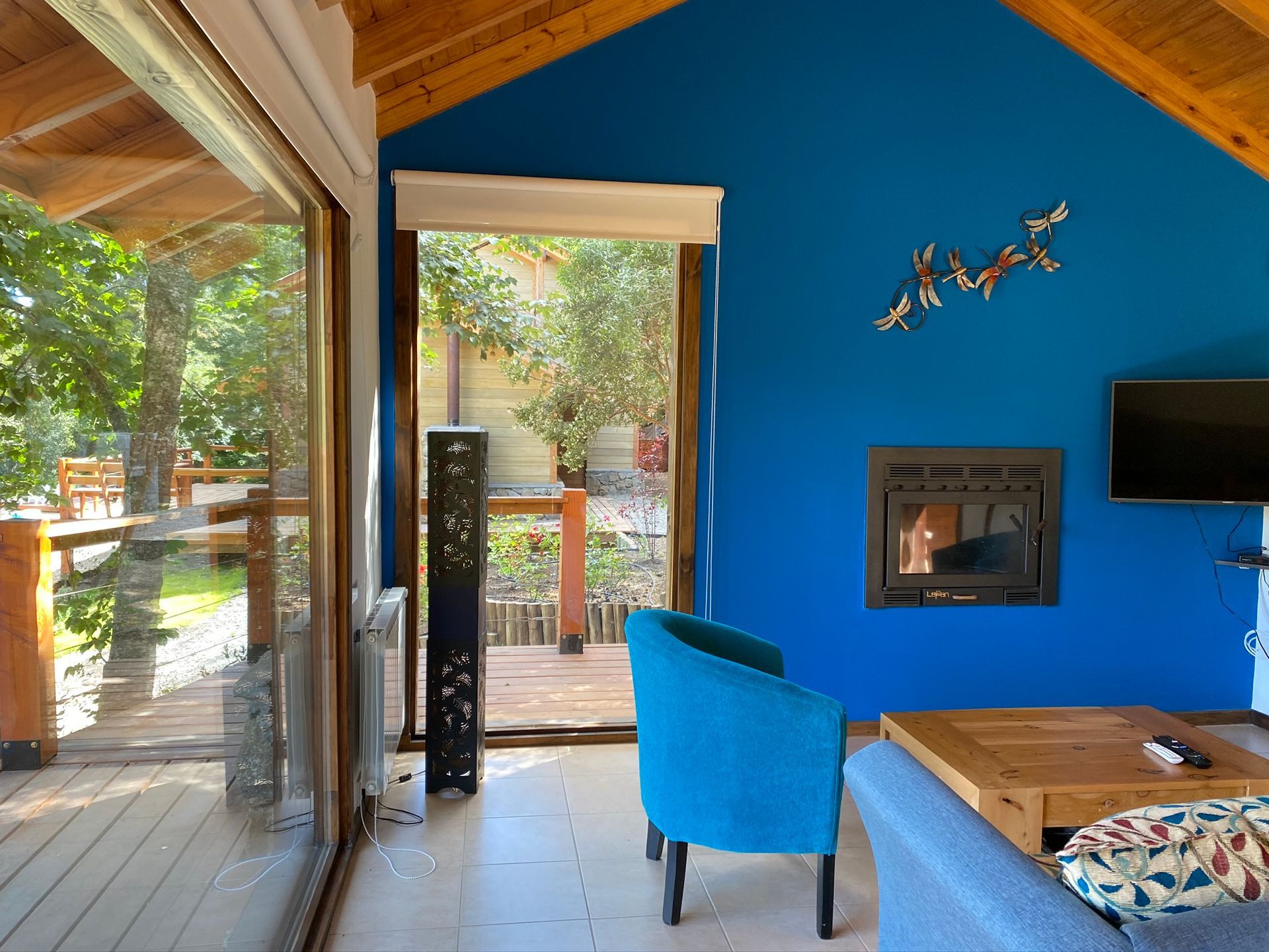 Una habitación con una pared de color azul brillante, una chimenea, un sillón azul, una mesa de madera y una puerta corredera que da a una terraza.