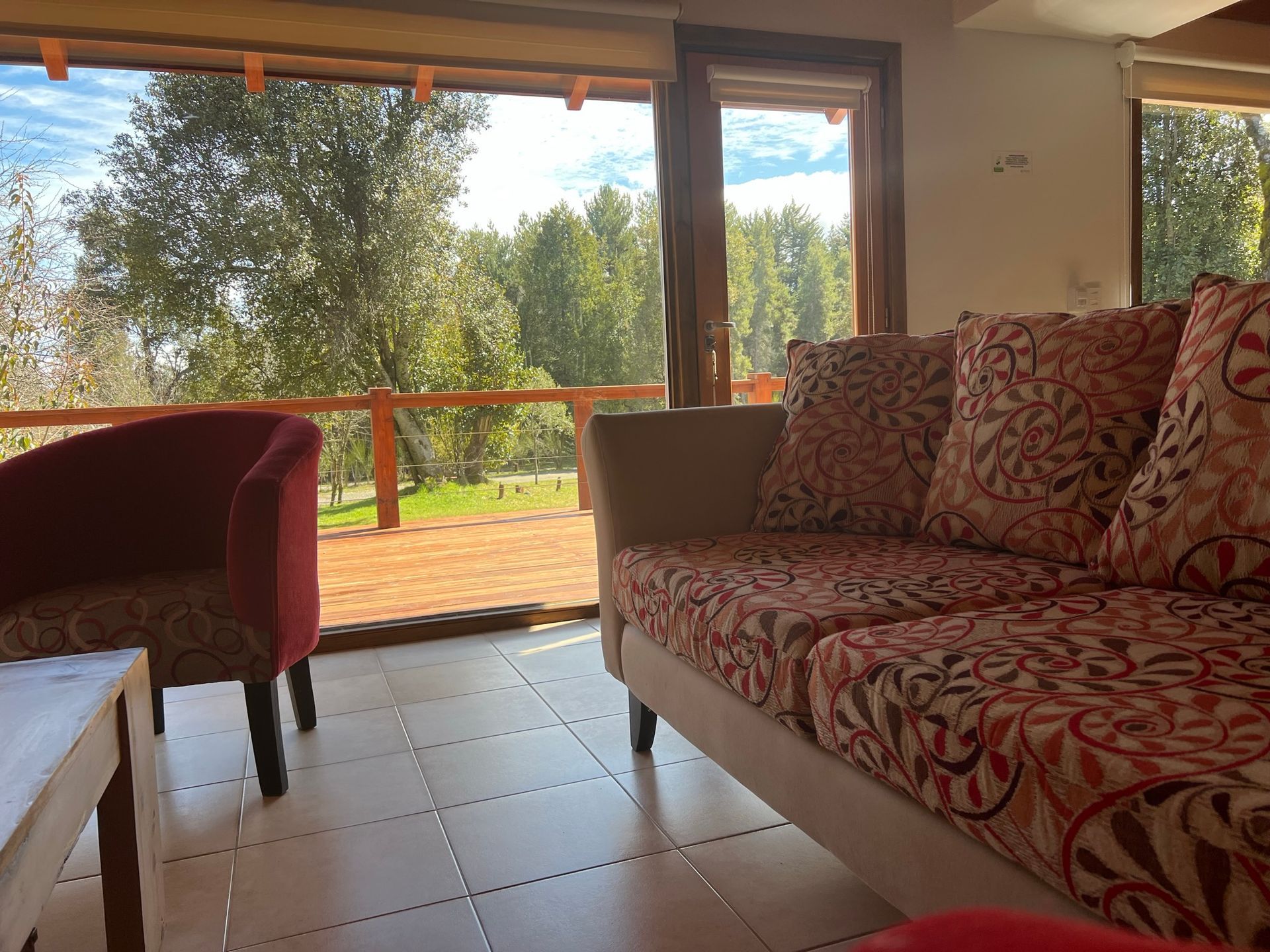 Sala de estar con un sofá tapizado con estampado, un sillón rojo y una gran ventana con vistas a los árboles y a una terraza de madera.