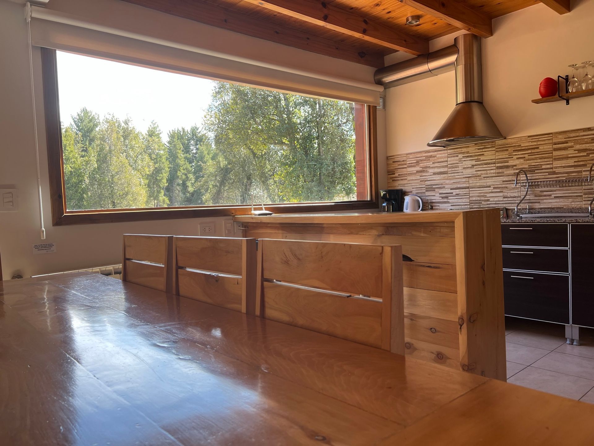 Una rústica isla de cocina de madera con taburetes se sitúa frente a una gran ventana con vistas a un paisaje luminoso y arbolado.
