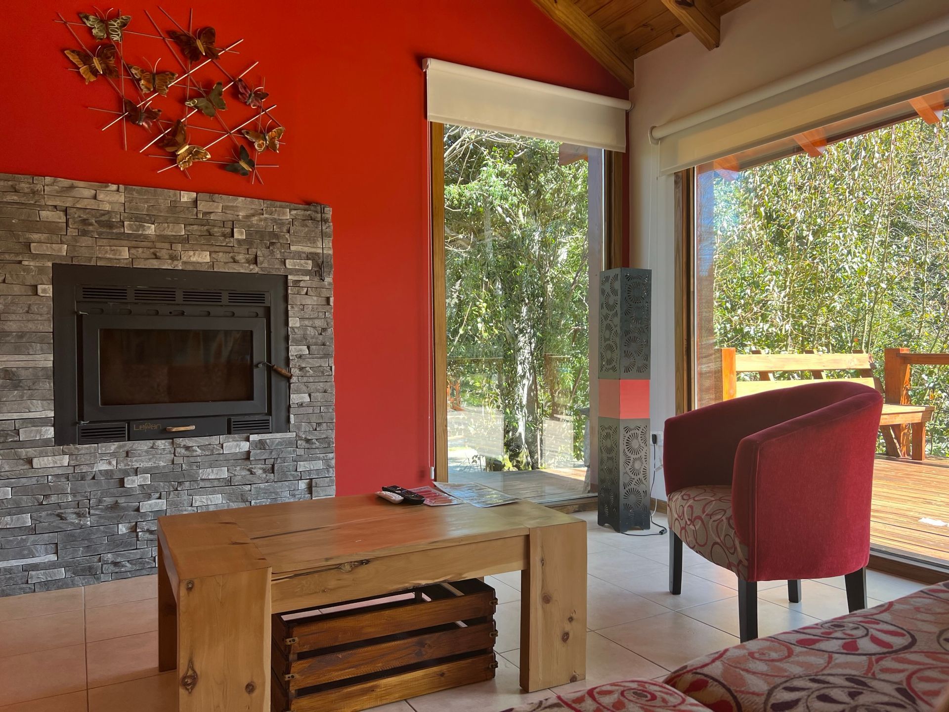 Una habitación con chimenea de piedra, pared roja decorada con mariposas, mesa de centro de madera, sillón rojo y grandes ventanales.