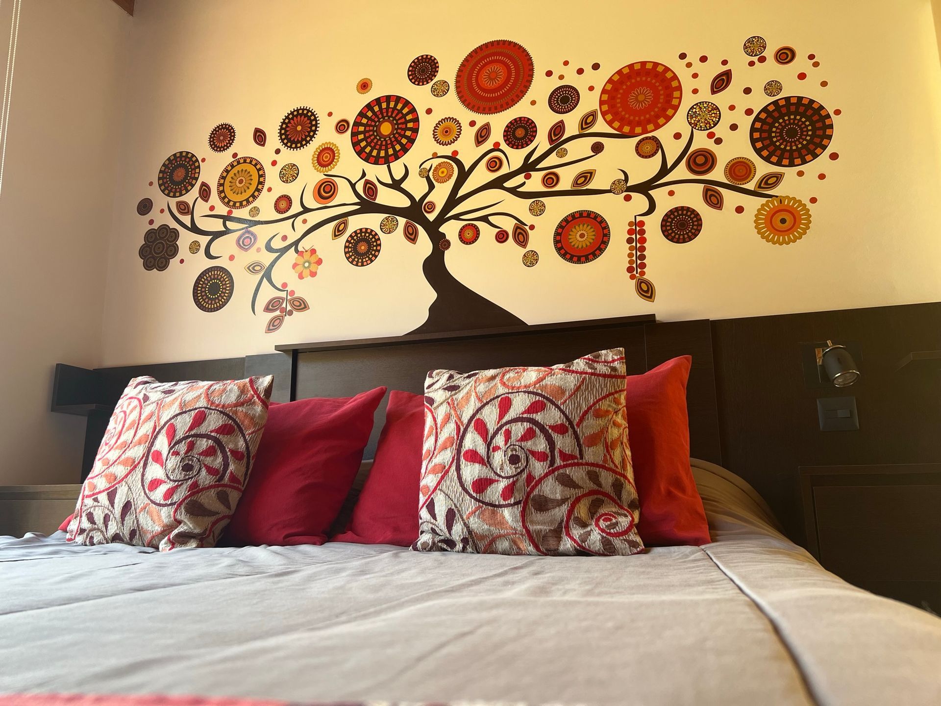 Una cama con cojines rojos y estampados se encuentra contra una pared que presenta un mural decorativo de un árbol estilizado con esferas florales.