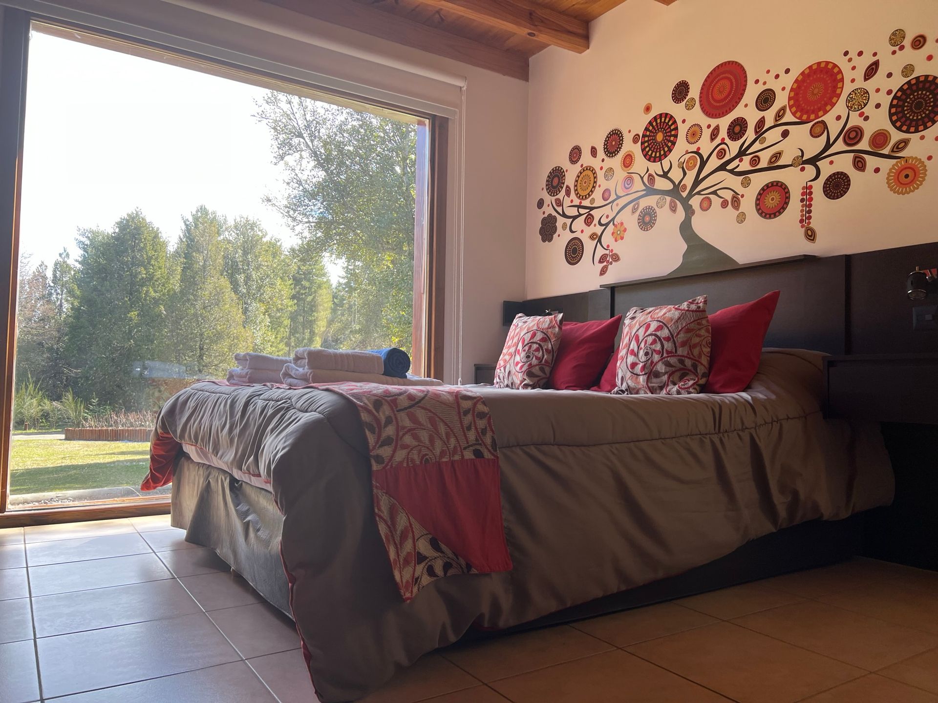 Un dormitorio moderno con cama, decoración floral roja en la pared y una gran ventana con vistas a un jardín soleado y arbolado.