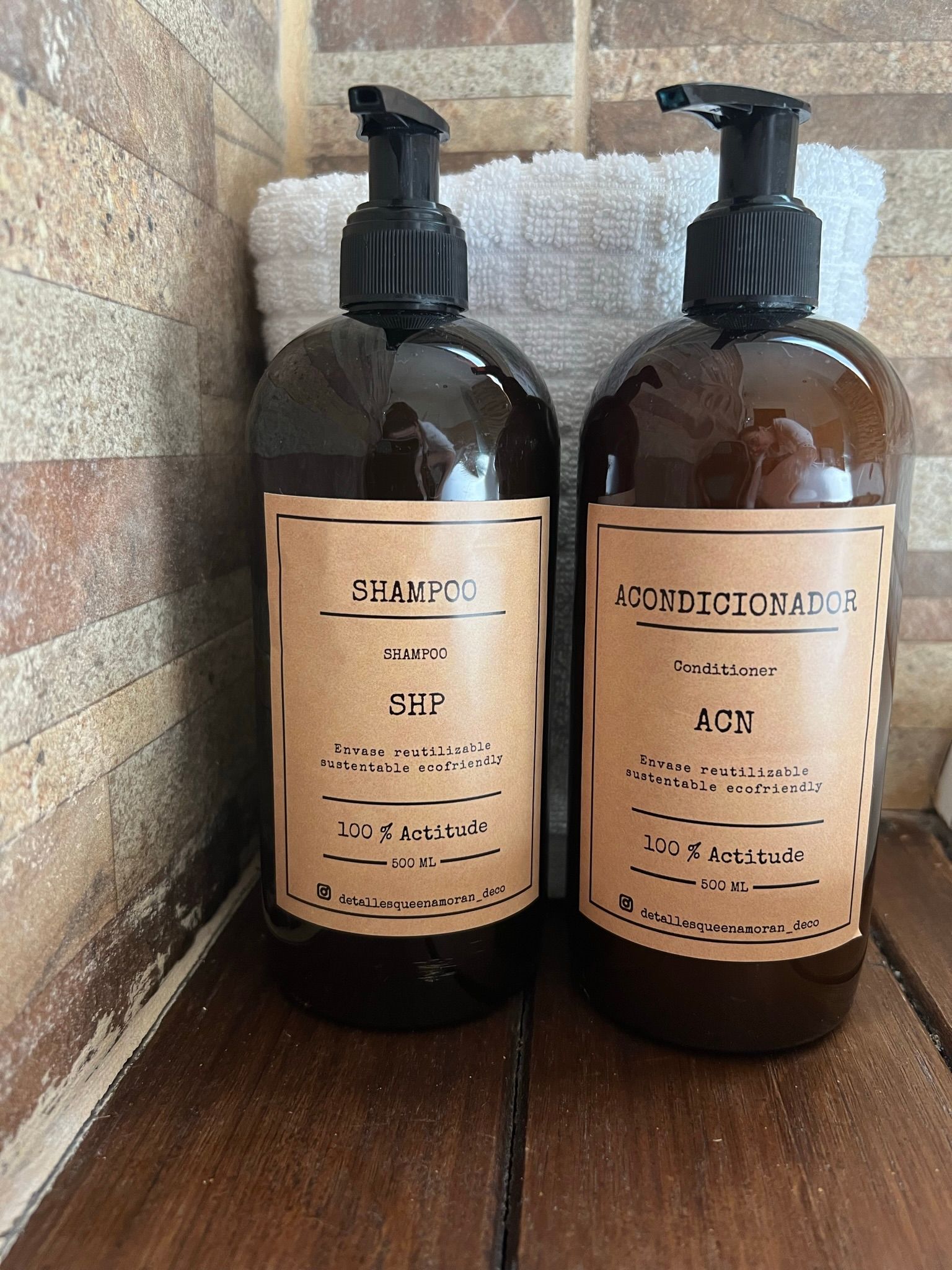 Dos frascos de color ámbar con dosificador y etiquetas minimalistas de color marrón reposan sobre una superficie de madera frente a una toalla blanca.