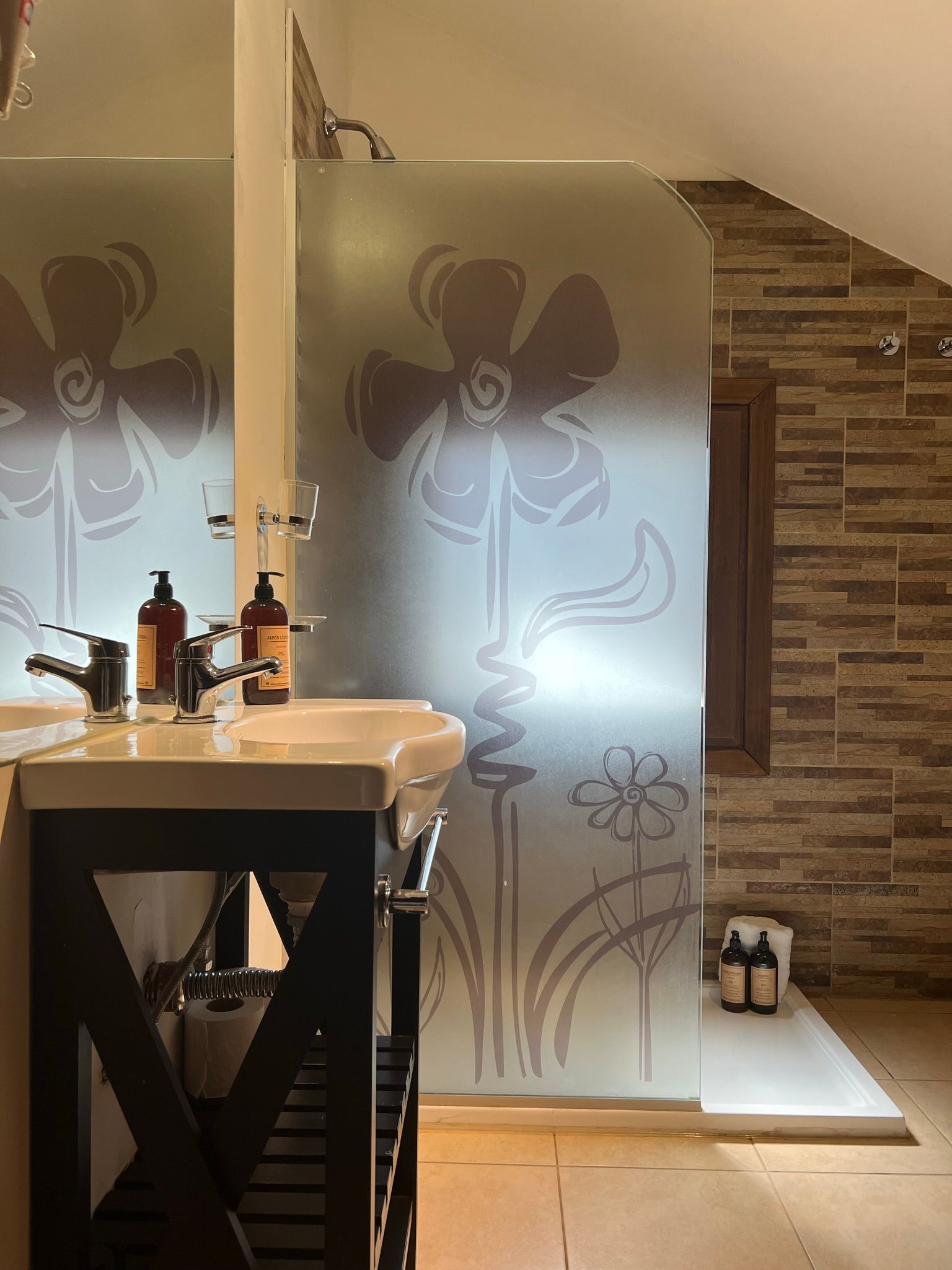 Un lavabo con grifo se sitúa junto a una puerta de ducha de cristal con diseño floral esmerilado y paredes revestidas de azulejos de piedra.