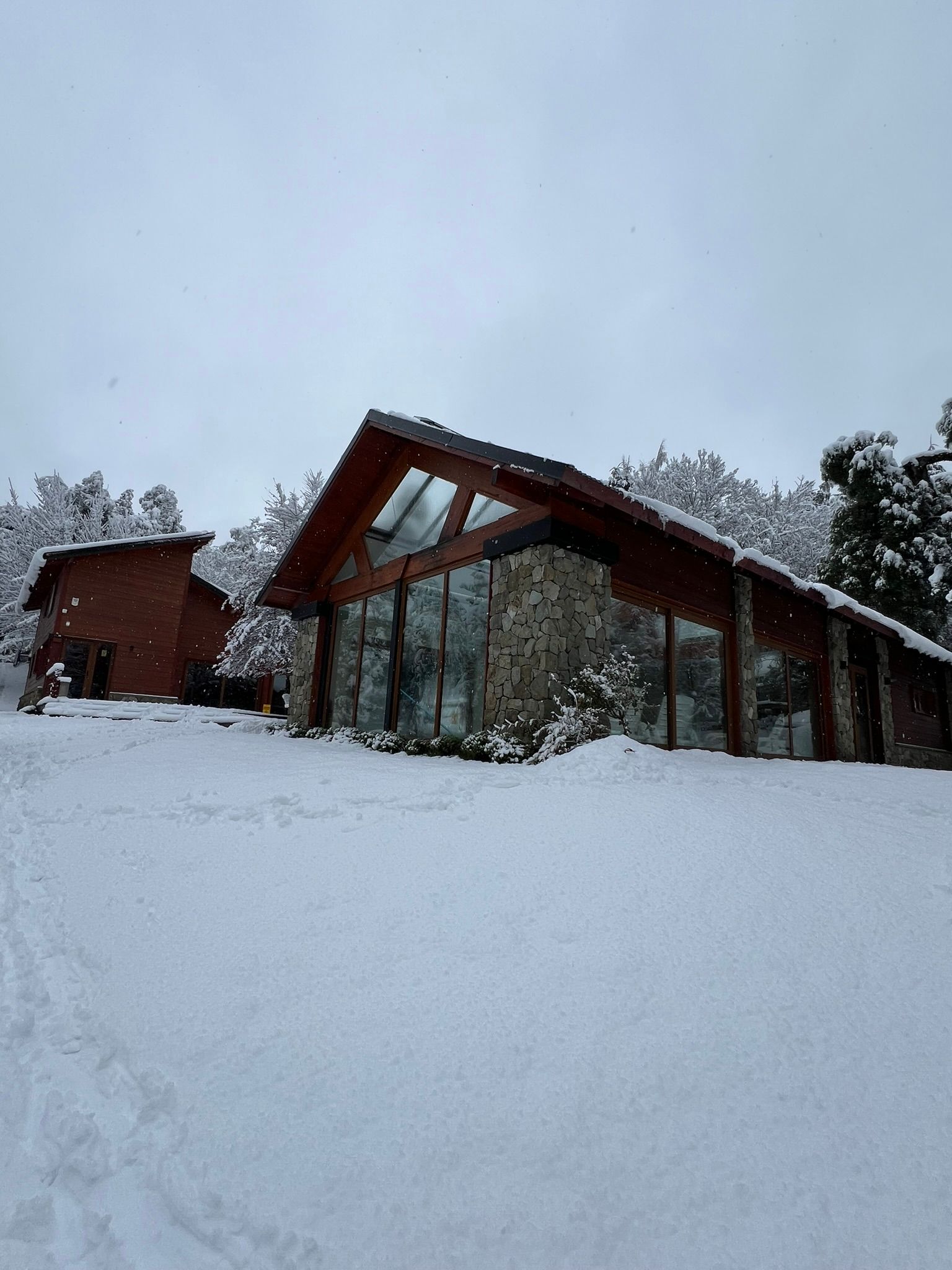 Dos cabañas modernas con fachada de piedra y estructura de madera se alzan en un paisaje nevado bajo un cielo invernal nublado.