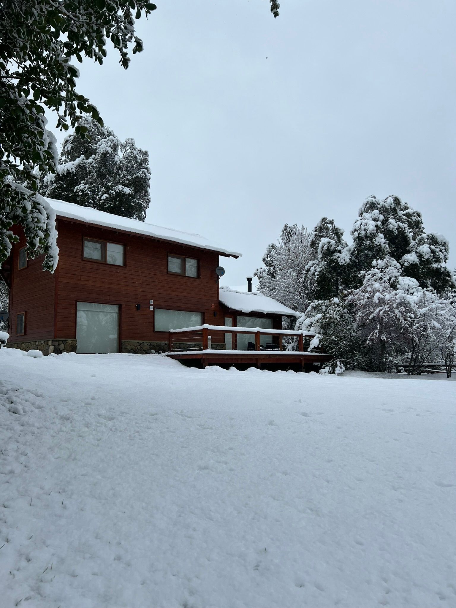Una casa de dos plantas de ladrillo rojo con una terraza de madera, situada en un patio cubierto de nieve blanca recién caída bajo un cielo nublado.