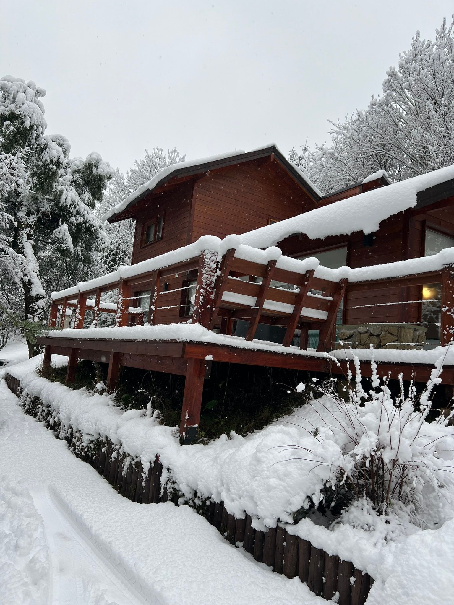 Una cabaña de madera con una terraza cubierta por una espesa capa de nieve blanca durante una nevada invernal, rodeada de árboles espolvoreados por la nieve.