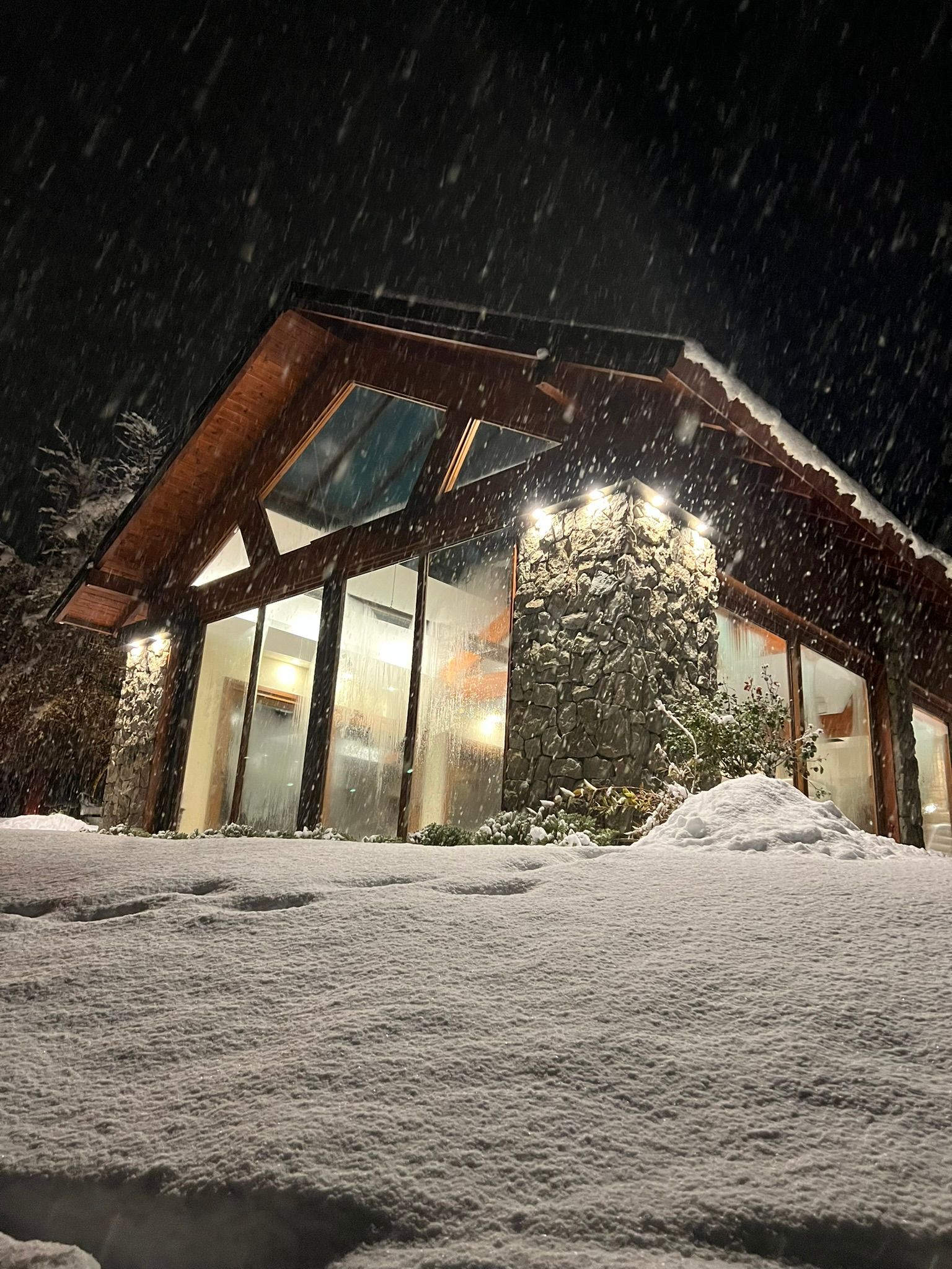 Una cabaña moderna e iluminada de noche durante una fuerte tormenta de nieve, con una gruesa capa de nieve cubriendo el suelo en primer plano.