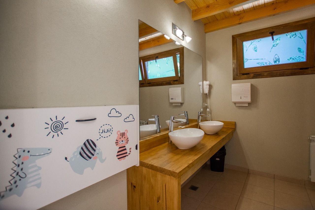 Un baño adaptado para niños, con dos lavabos blancos sobre una encimera de madera, un espejo y un vinilo decorativo de animales en la pared.