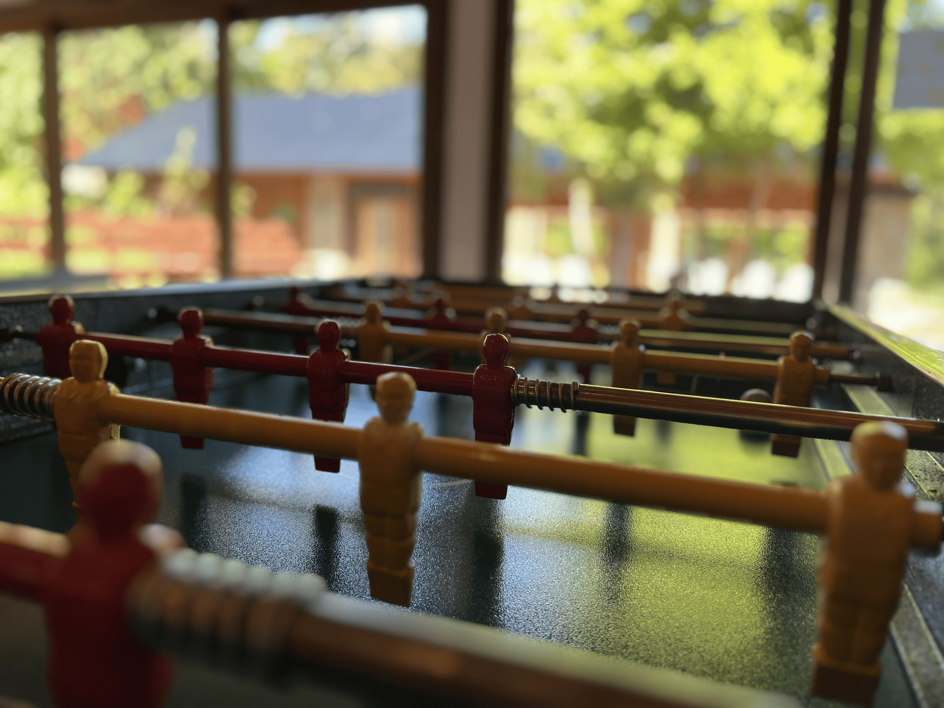 Primer plano de una mesa de futbolín con figuras de jugadores rojas y beige, situada en una habitación luminosa con grandes ventanales.