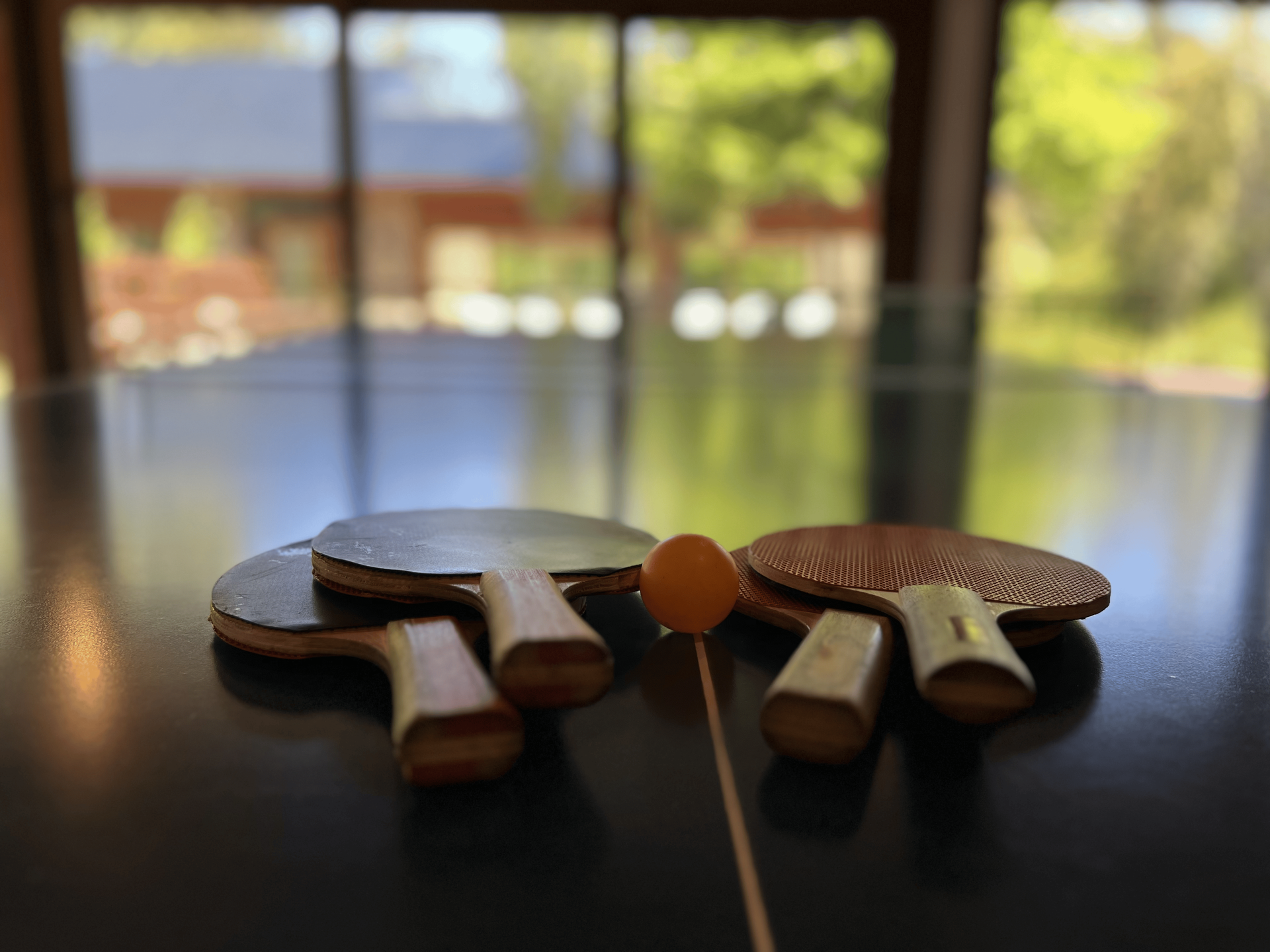 Cuatro palas de tenis de mesa de madera y una pelota amarilla colocadas sobre una mesa de ping-pong negra con un fondo exterior borroso.