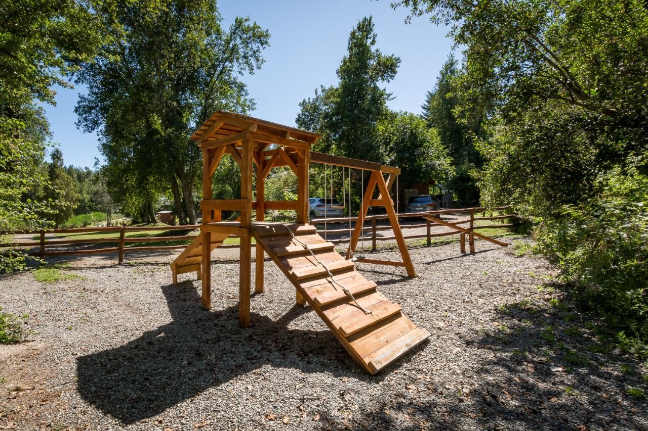 Estructura de juegos de madera con rampa para escalar y columpios sobre una superficie de virutas de madera, rodeada de árboles.