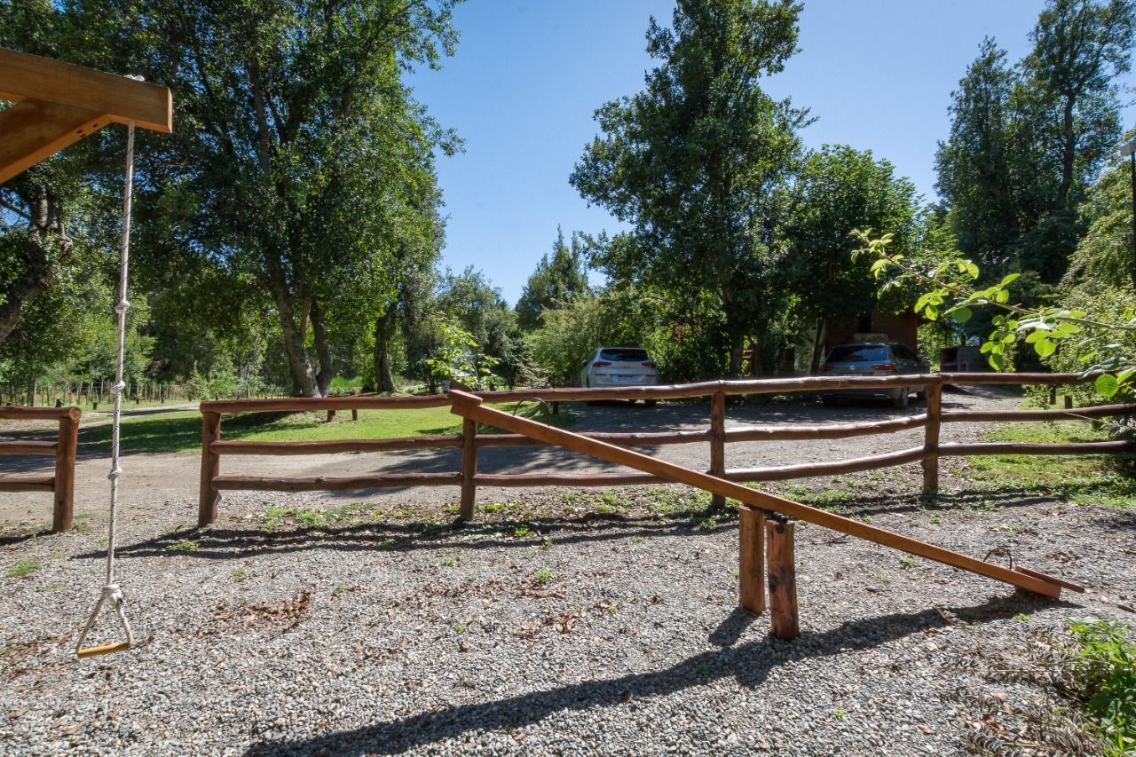 Un balancín y un columpio en un parque infantil de grava, frente a una valla de madera, árboles y coches aparcados bajo un cielo azul.