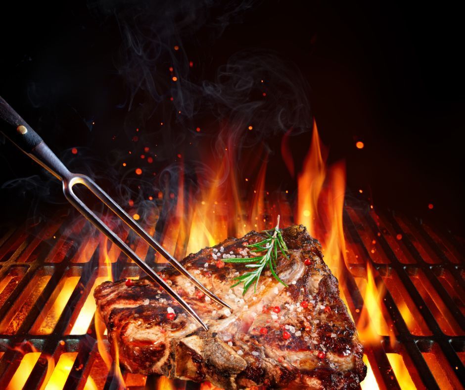 Un filete T-bone asándose a la parrilla sobre fuego abierto, sazonado con hierbas y pimiento rojo, y atravesado por un tenedor de carne de metal.
