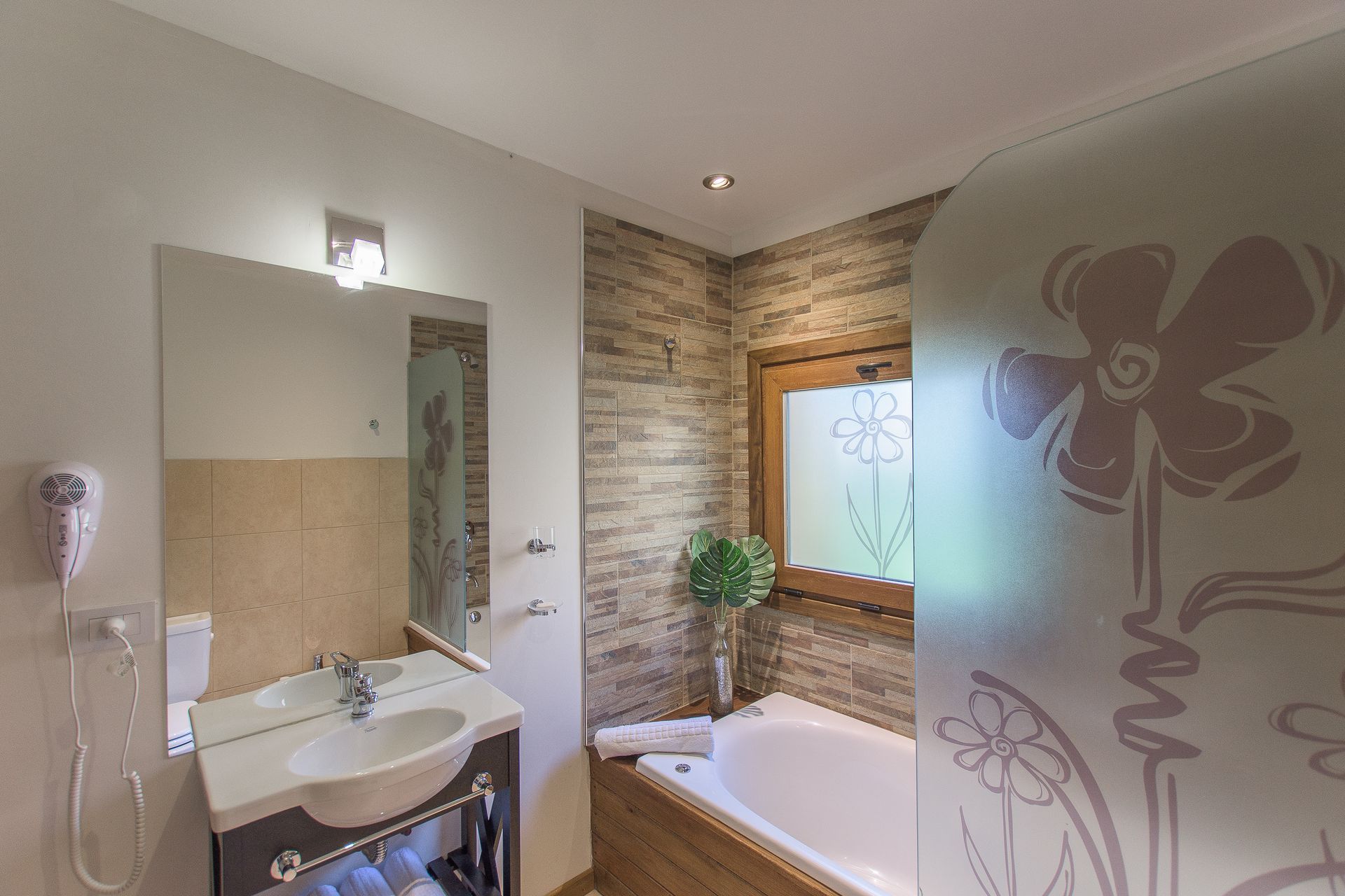 Un baño moderno con lavabo blanco, espejo, bañera, paredes con textura de piedra y puerta de cristal esmerilado con motivos florales.